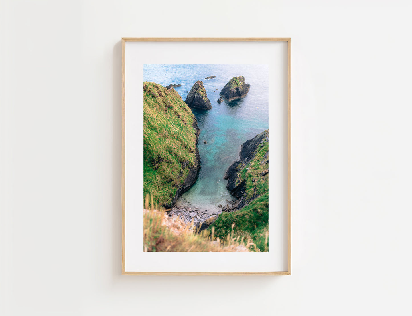 A framed coastal landscape photograph hanging on a white wall. The print shows turquoise water, green cliffs, and dark rock formations at Dun Chaoin in Ireland, surrounded by a white mat and a light natural wood frame.
