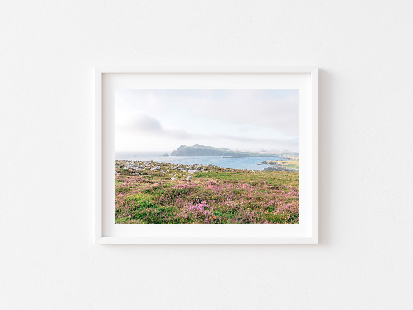 A framed landscape photograph of a coastal field with pink heather, distant cliffs at Ceann SIbeal, and a cloudy sky, displayed on a clean white wall.