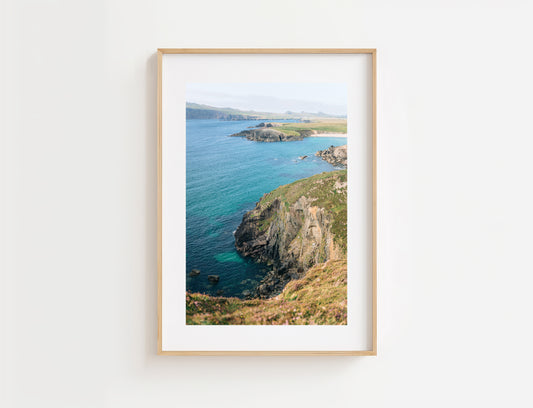 A framed coastal landscape photograph hanging on a white wall. The print shows turquoise ocean water, rocky cliffs of Ceann Sibeal in Ireland, and distant green hills under a pale sky, surrounded by a white mat and a light natural wood frame.