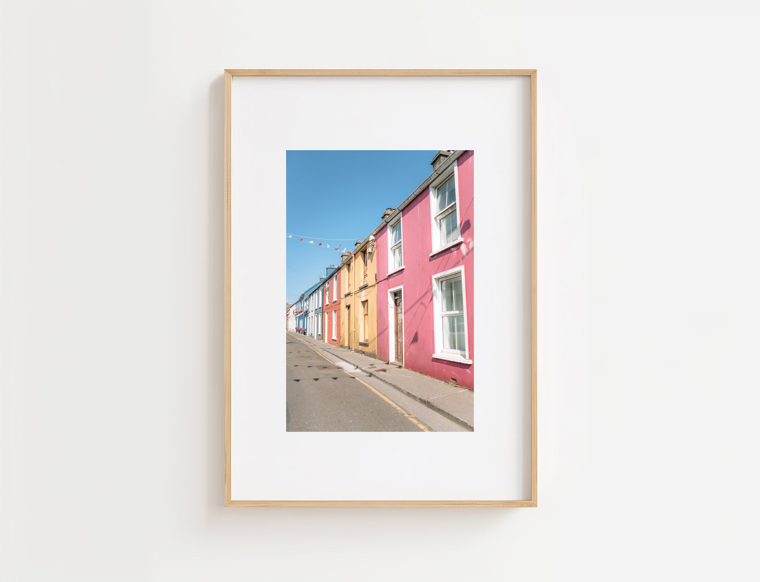 A framed art print featuring a photograph of colorful row houses lining a street under a clear blue sky, displayed on a clean white wall.