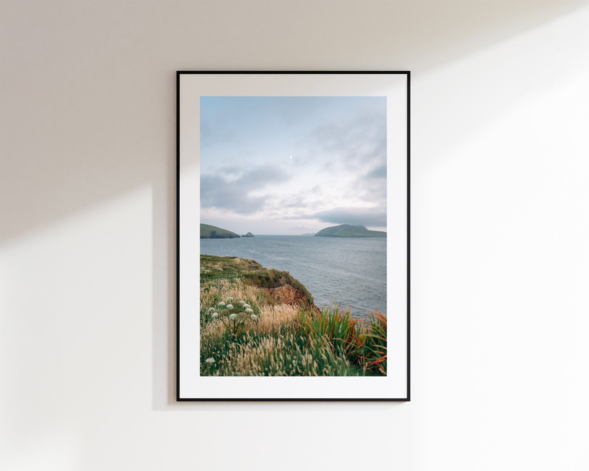A framed coastal landscape print hangs on a white wall. The artwork shows a cloudy sky over calm water with distant islands and grassy cliffs of Dun Chaoin in the foreground.