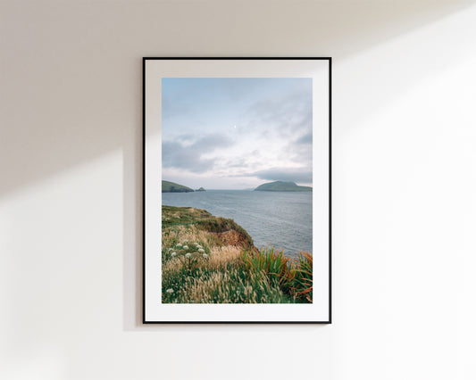 A framed coastal landscape print hangs on a white wall. The artwork shows a cloudy sky over calm water with distant islands and grassy cliffs of Dun Chaoin in the foreground.