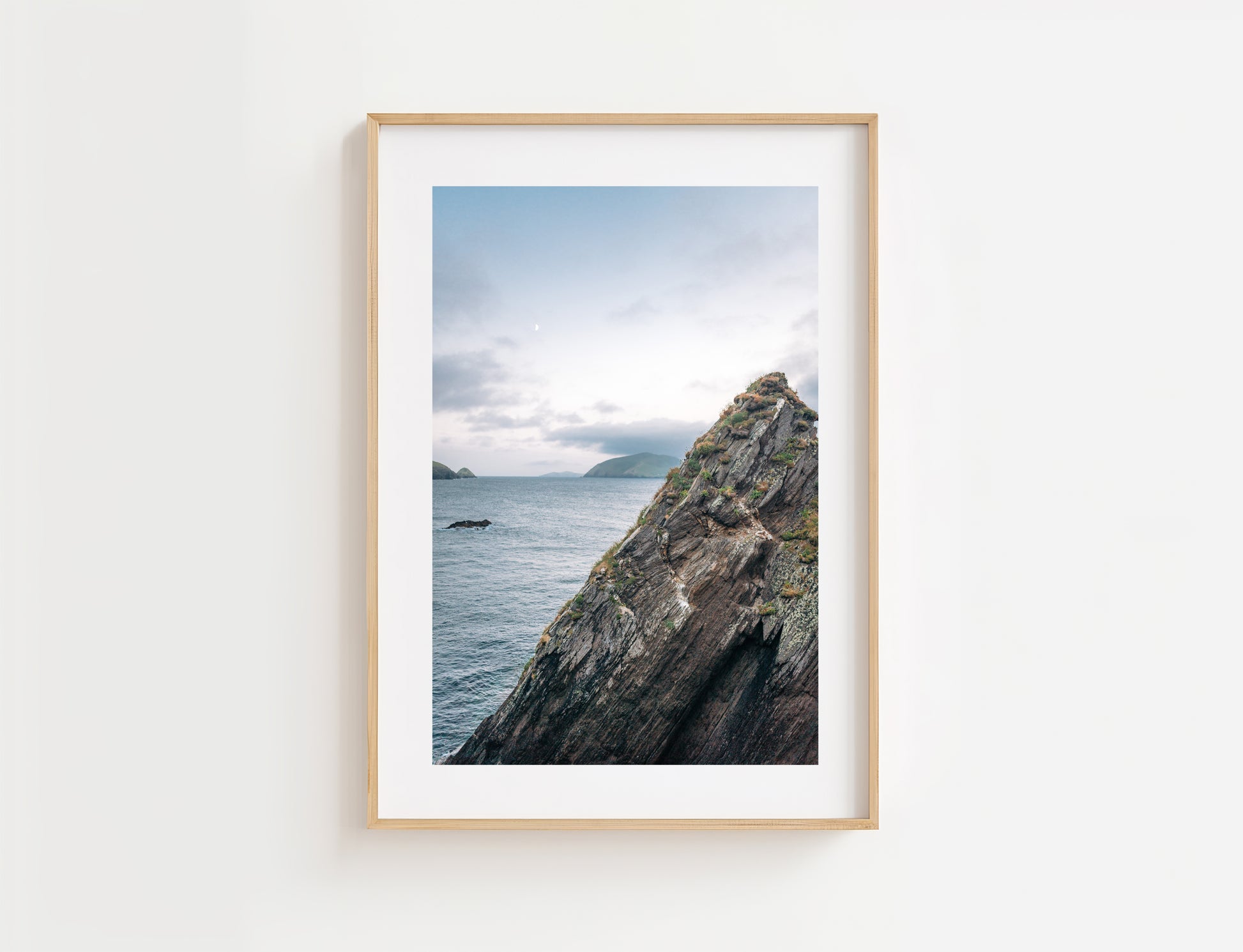 A framed photographic print of the rugged rocky coastline of Dun Chaoin over a calm sea under a cloudy sky, displayed on a clean white wall.