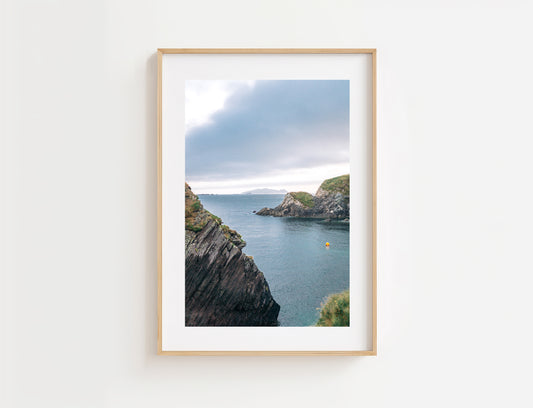 A framed landscape art print depicting a coastal scene with rocky cliffs and blue-gray water at Dun Chaoin under a cloudy sky, displayed on a clean white wall.