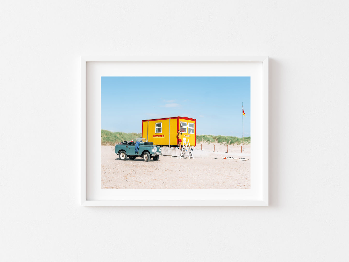 A framed art print hanging on a white wall, depicting a beach scene with a lifeguard on duty in front of a small yellow and red booth, a blue ford bronco, sand dunes, and a clear blue sky.