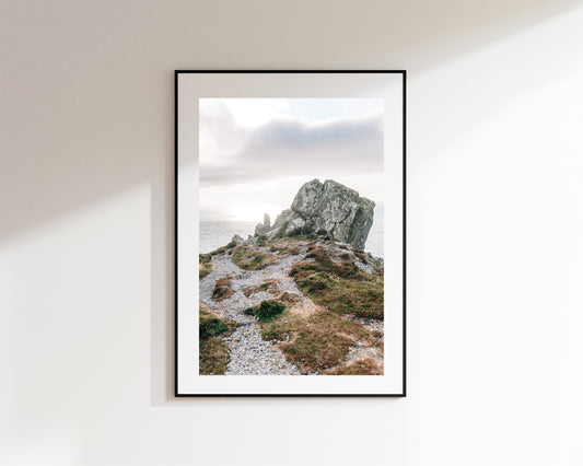 A framed landscape art print depicting a rocky coastal outcrop at Ceann Sibeal under a cloudy sky, mounted on a white wall.